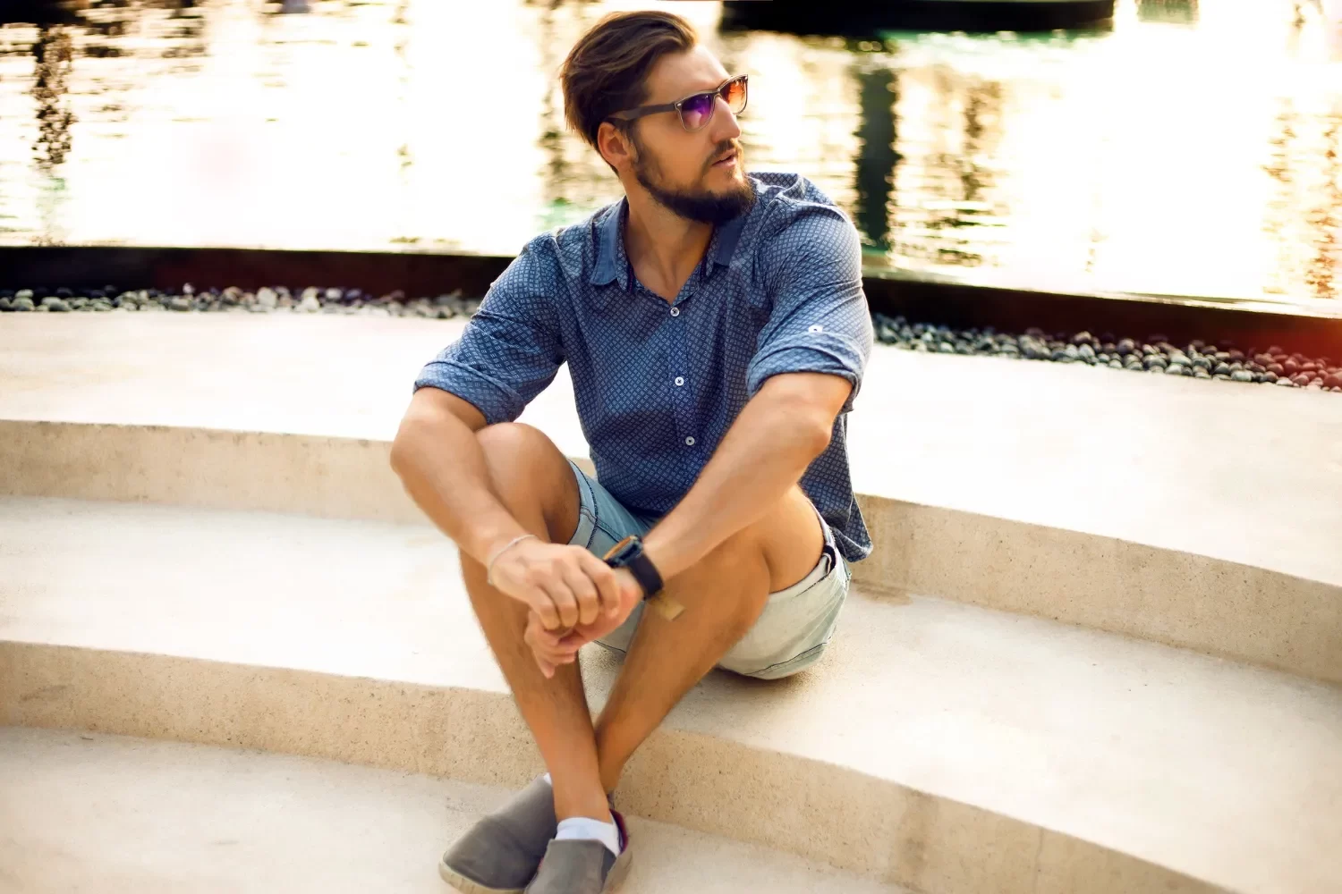 Hombre sentado en escaleras de concreto junto a un estanque, vistiendo una camisa de cuadros y pantalones cortos, con gafas de sol y reloj en la muñeca.