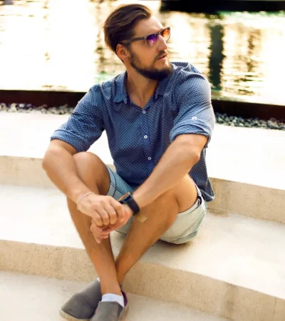 Hombre sentado en escaleras de concreto junto a un estanque, vistiendo una camisa de cuadros y pantalones cortos, con gafas de sol y reloj en la muñeca.