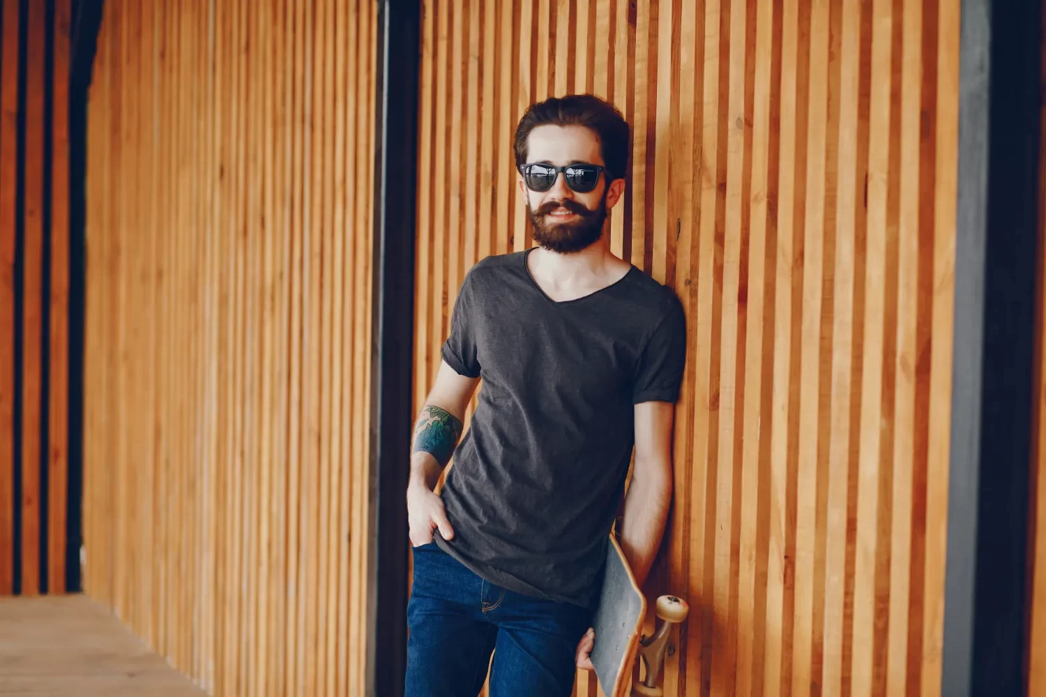 Hombre con barba y gafas de sol apoyado en una pared de madera, vistiendo una camiseta oscura y jeans, sosteniendo una patineta.