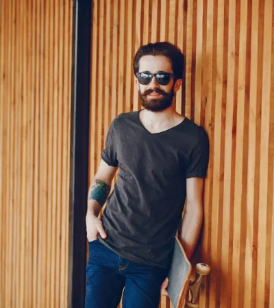 Hombre con barba y gafas de sol apoyado en una pared de madera, vistiendo una camiseta oscura y jeans, sosteniendo una patineta.