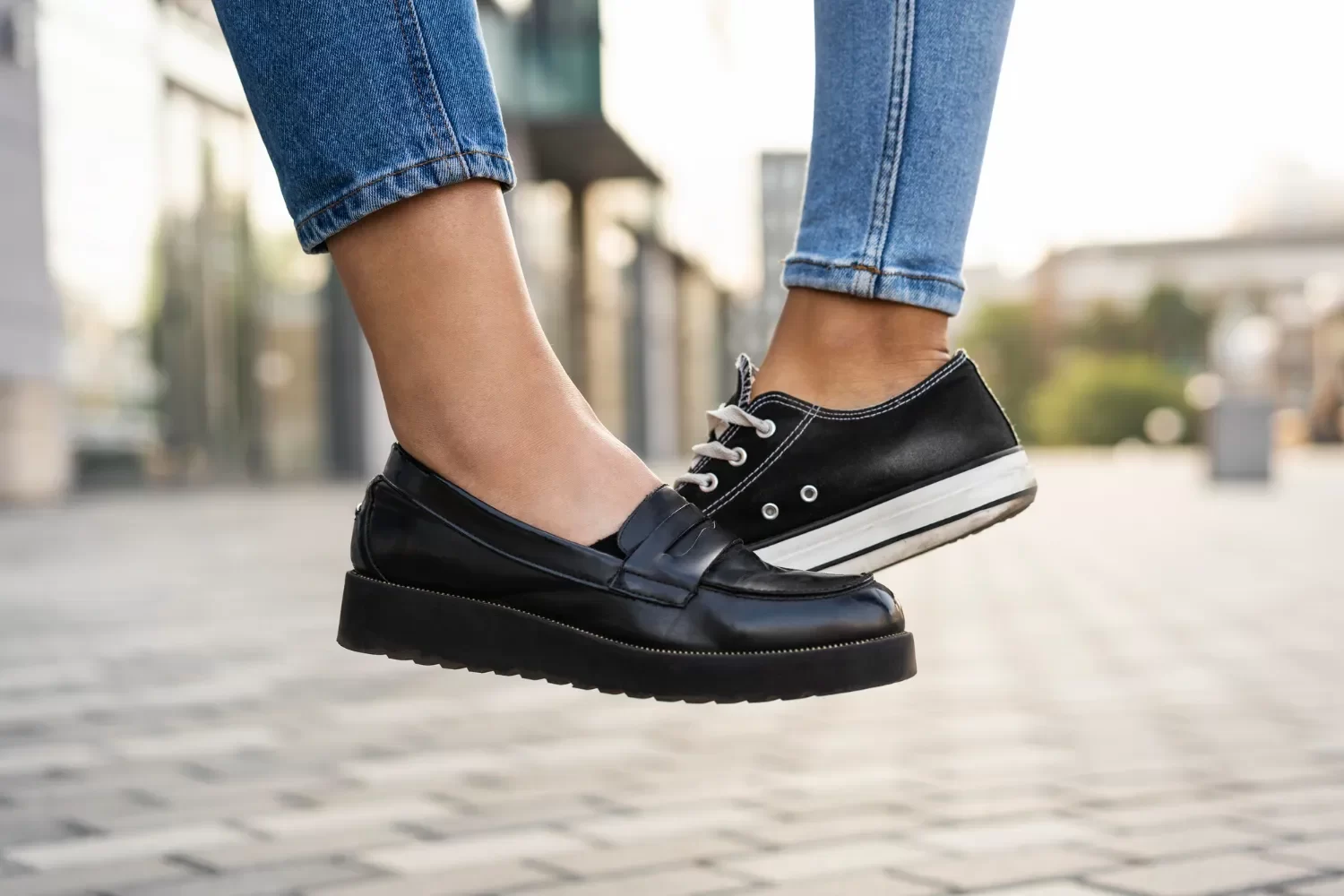 Dos zapatos en primer plano, uno negro tipo mocasín y otro negro deportivo con suela blanca, ambos llevados por una persona de pie sobre una superficie pavimentada.