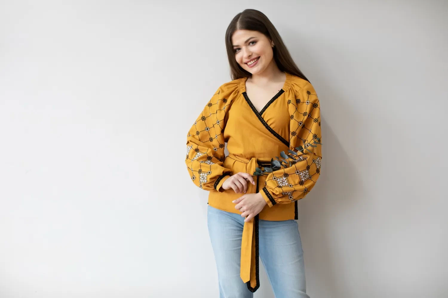 Mujer sonriente con blusa amarilla de manga larga y detalles bordados, sosteniendo una ramita de eucalipto, posando frente a un fondo blanco.