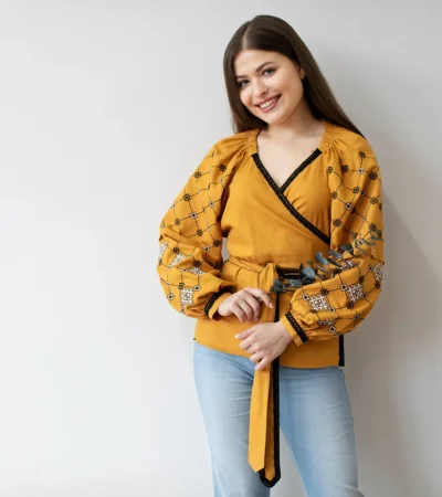 Mujer sonriente con blusa amarilla de manga larga y detalles bordados, sosteniendo una ramita de eucalipto, posando frente a un fondo blanco.